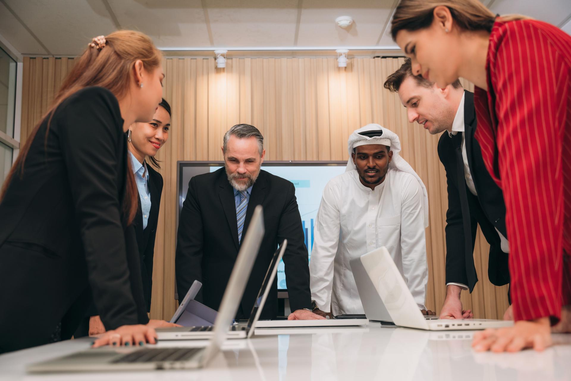 A group of different business people are sitting around a big meeting table in a modern office.